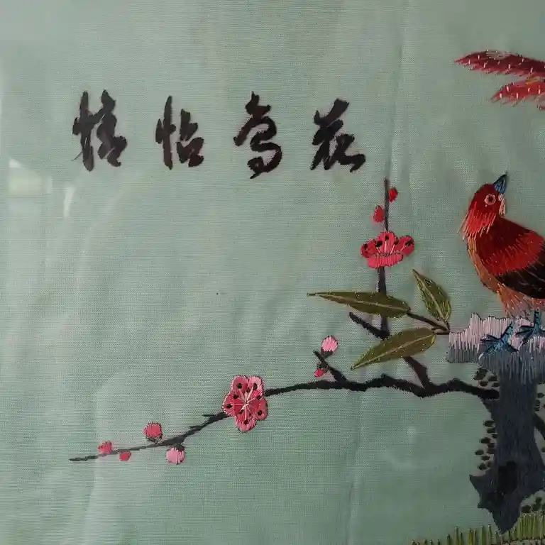 A close-up of traditional Chinese embroidery on light green fabric, featuring a red bird perched on a blossoming branch with…
