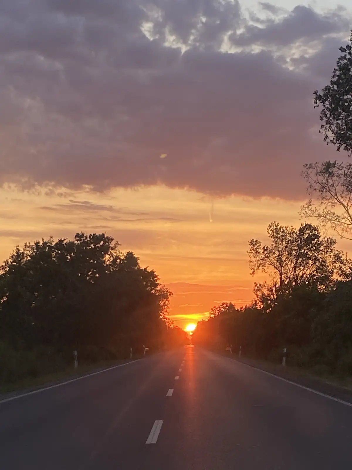 A scenic photo of a road stretching toward a vibrant sunset with orange and purple clouds, flanked by trees. The overlaid…