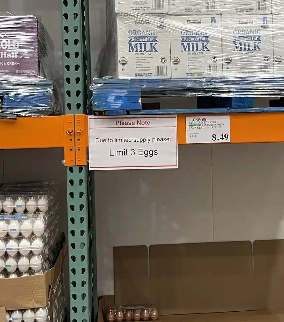 A photo of a Costco shelf with egg cartons on the lower level and milk cases on the upper level, featuring a white sign that…