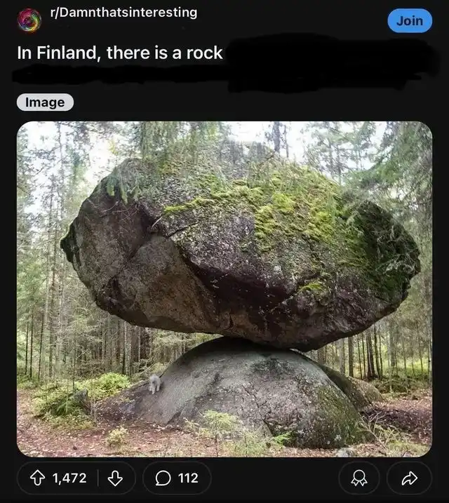 A large, moss-covered boulder is precariously balanced on top of a smaller, rounded boulder in a dense forest. The image is…