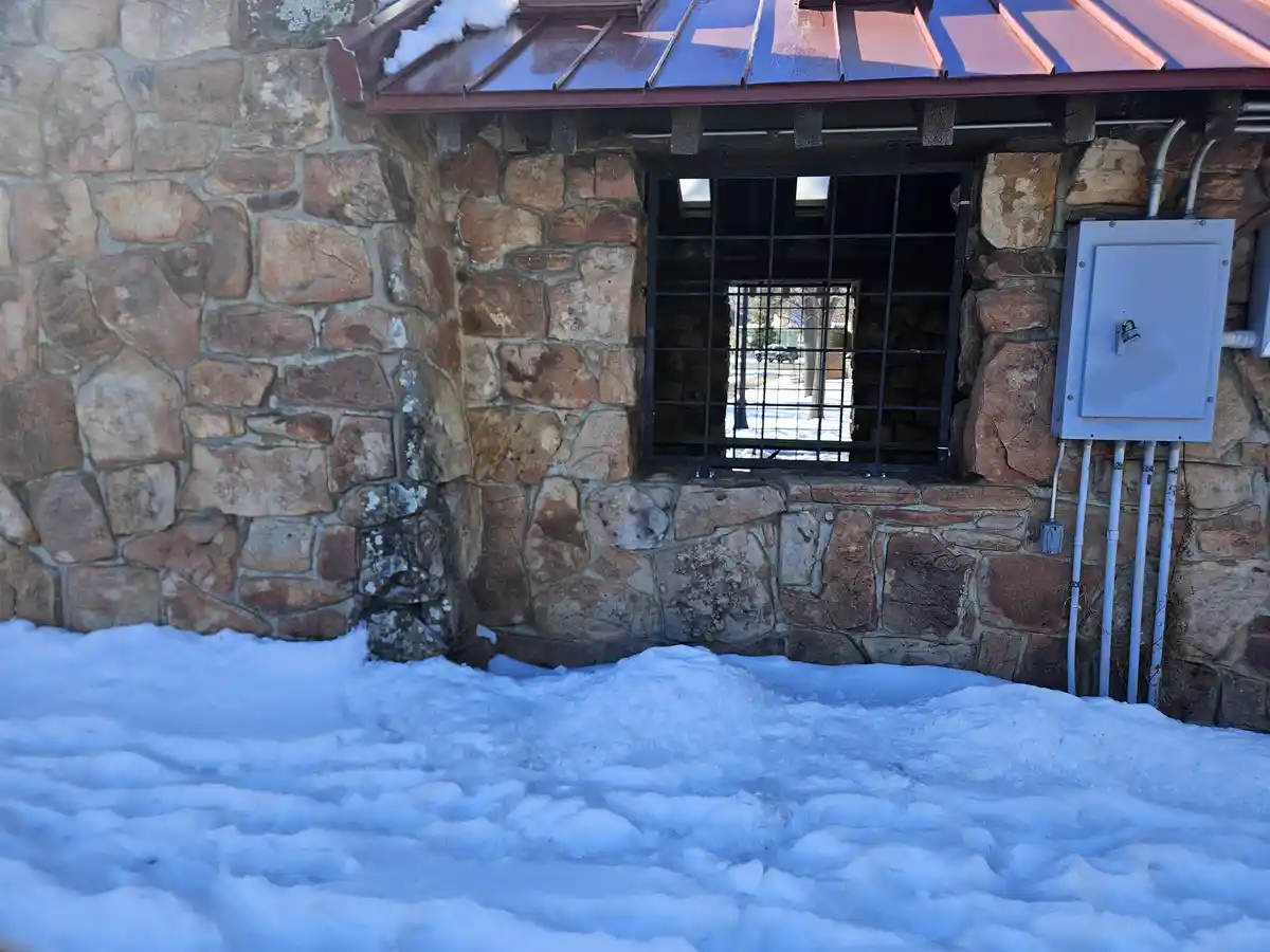 A snowy outdoor shot of a stone building with a barred window and a locked electrical box. The ground is covered in deep…
