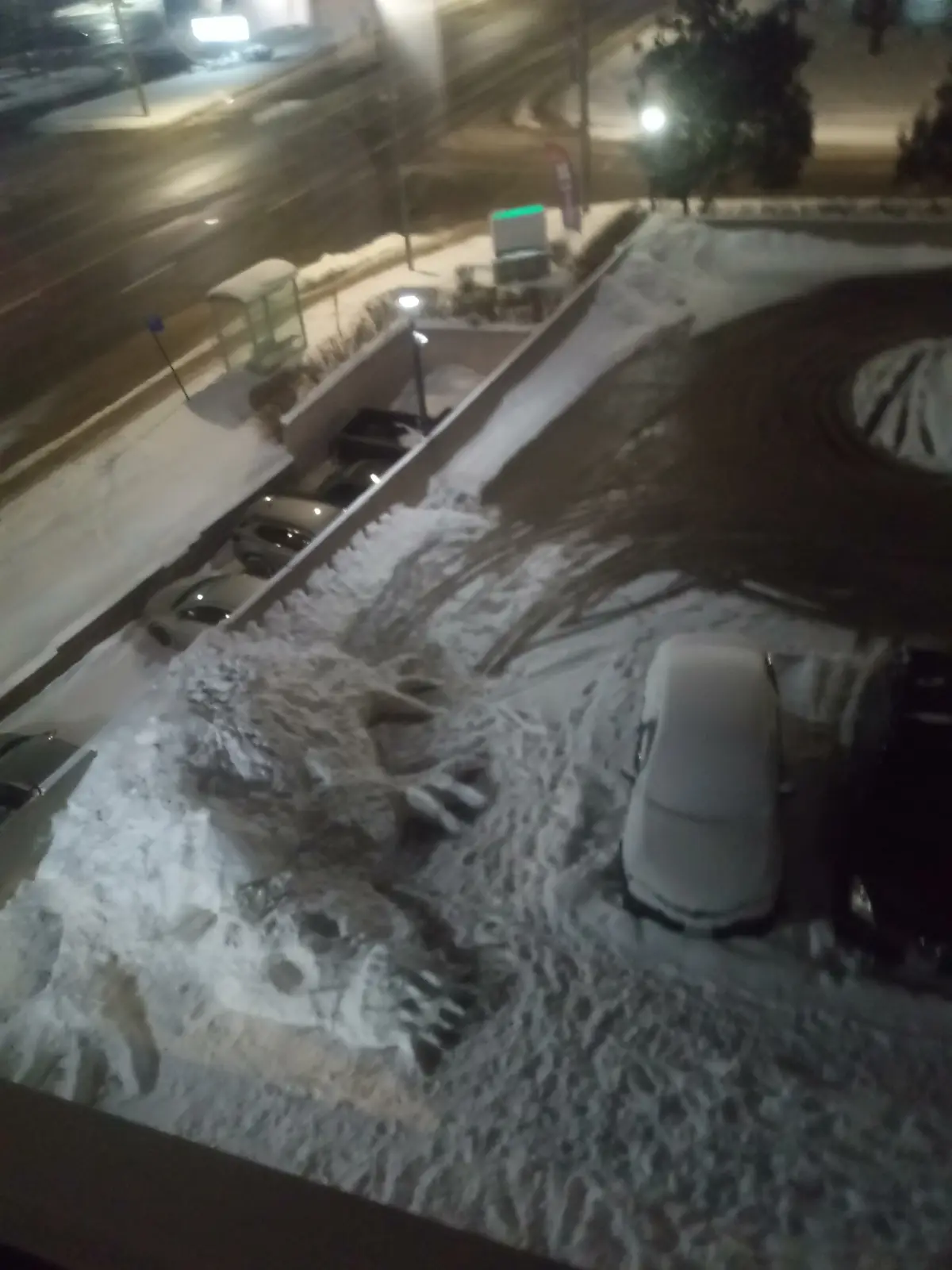 A nighttime aerial view of a snowy parking lot where snow has been piled and shaped into a large, monstrous creature with…