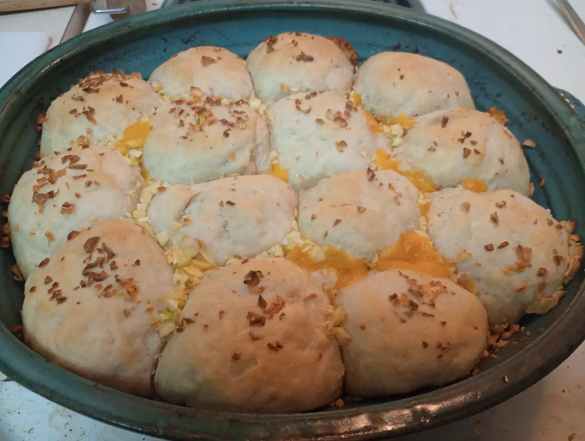A close-up photo shows a green ceramic baking dish filled with golden-brown, round baked rolls, some split open to reveal…