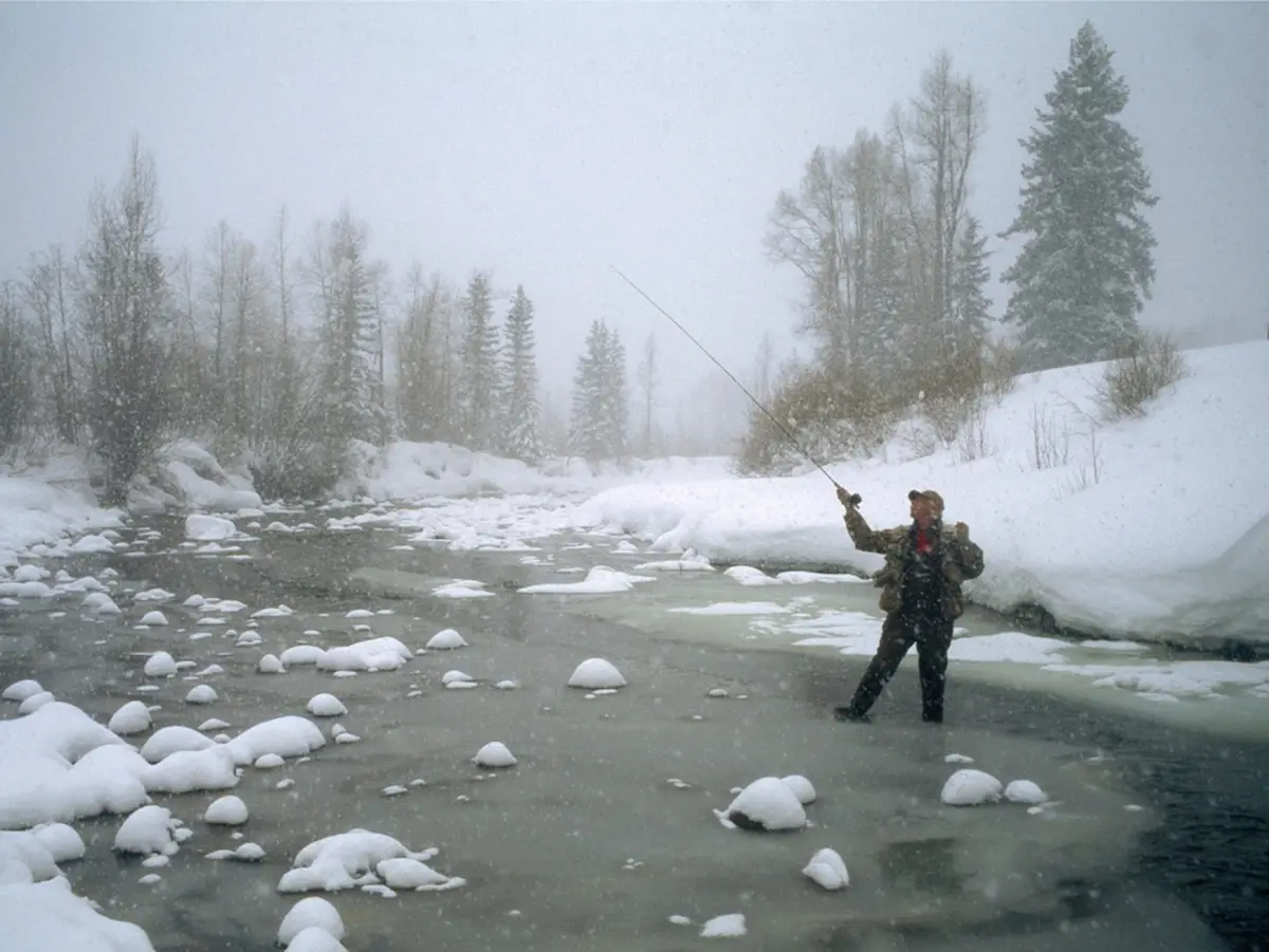 A man stands in a snow-covered, partially frozen river, casting a fly fishing rod while snow falls heavily around him. He…