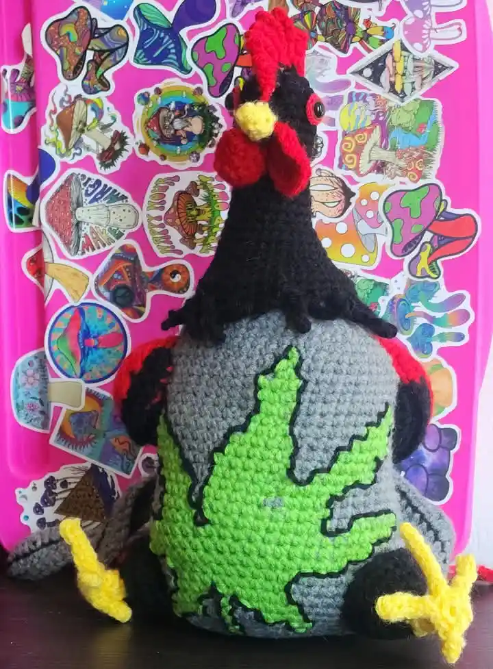 A crocheted chicken plushie with a bright green pine tree stitched onto its grey back sits atop a pink surface covered in…