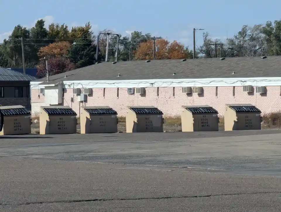 A row of beige dumpsters in front of a pink brick building, each labeled 'NO ROCK WOOD METAL', with one dumpster's text…