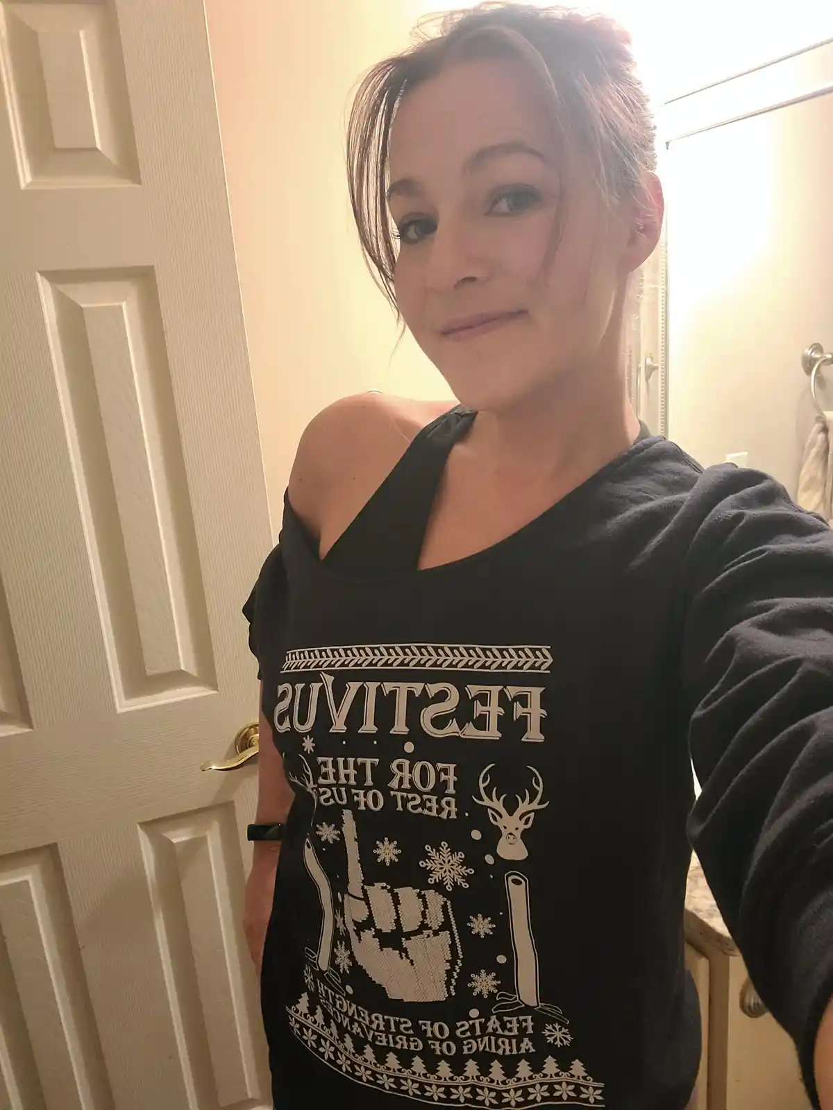A woman takes a selfie in a bathroom wearing a black long-sleeved shirt with white text and graphics. The shirt reads…