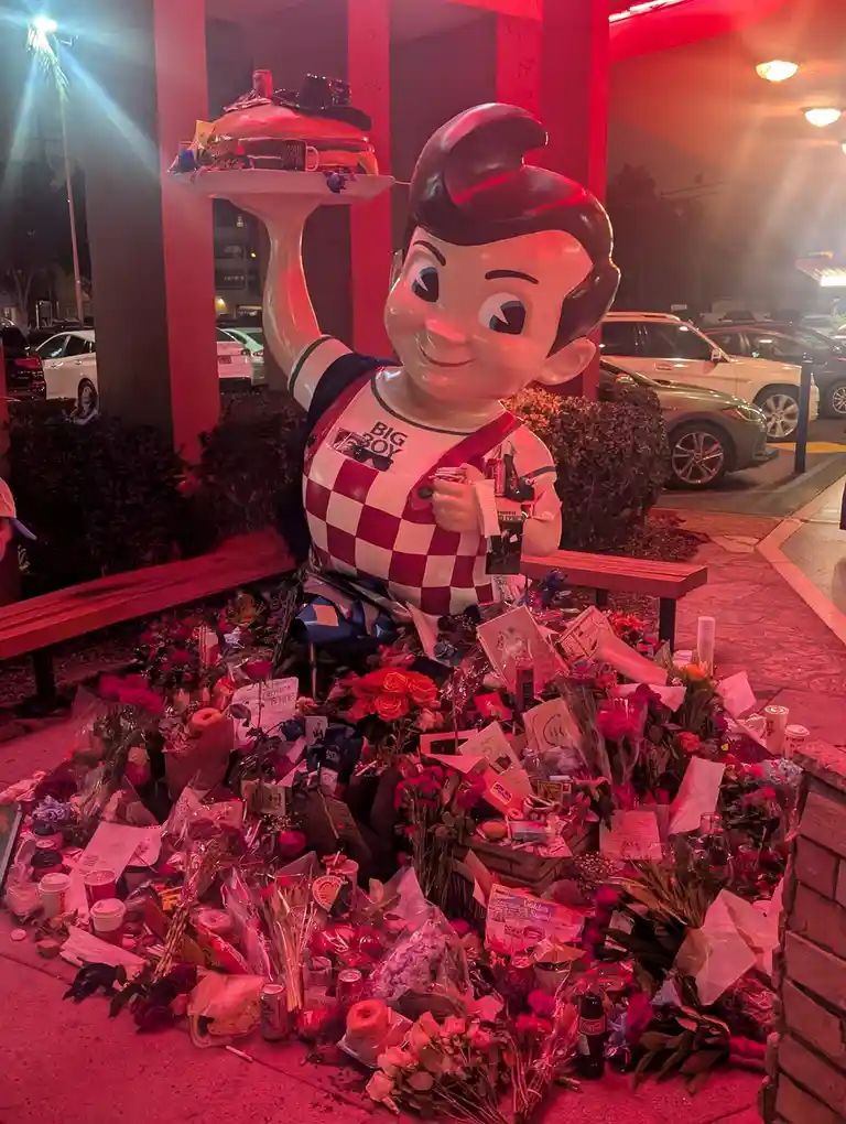 A large, smiling Big Boy statue holding a tray of food stands at night, surrounded by a massive pile of flowers, handwritten…