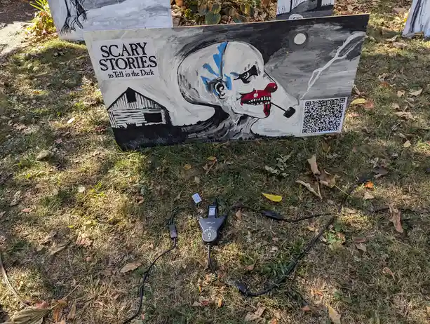 A hand-painted sign outdoors depicts a menacing clown with blue face paint, red lips, and a pipe, set against a dark sky…