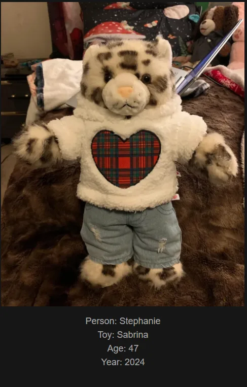A plush tiger toy named Sabrina, dressed in a white fuzzy hoodie with a red plaid heart on the front and light blue denim…