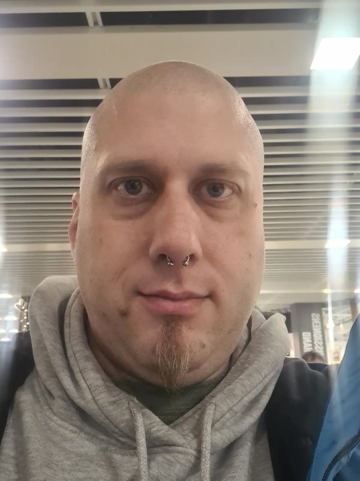 A bald man with a nose ring and goatee takes a selfie indoors, wearing a grey hoodie and a blue jacket. The overlaid text…