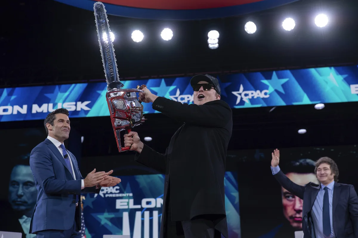 A man in a black coat and sunglasses holds up a large chainsaw on a stage with 'cpac' branding, while two other men in suits…