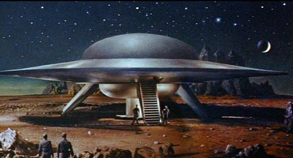 A vintage sci-fi movie still shows a large, silver, saucer-shaped UFO landed on a rocky, alien planet under a starry sky…