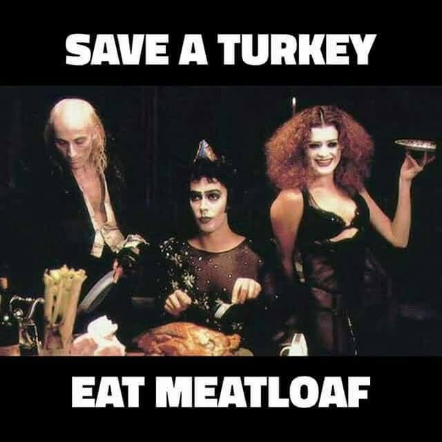 Three women in dark, theatrical costumes are gathered around a roasted turkey on a table. The woman on the left holds a…