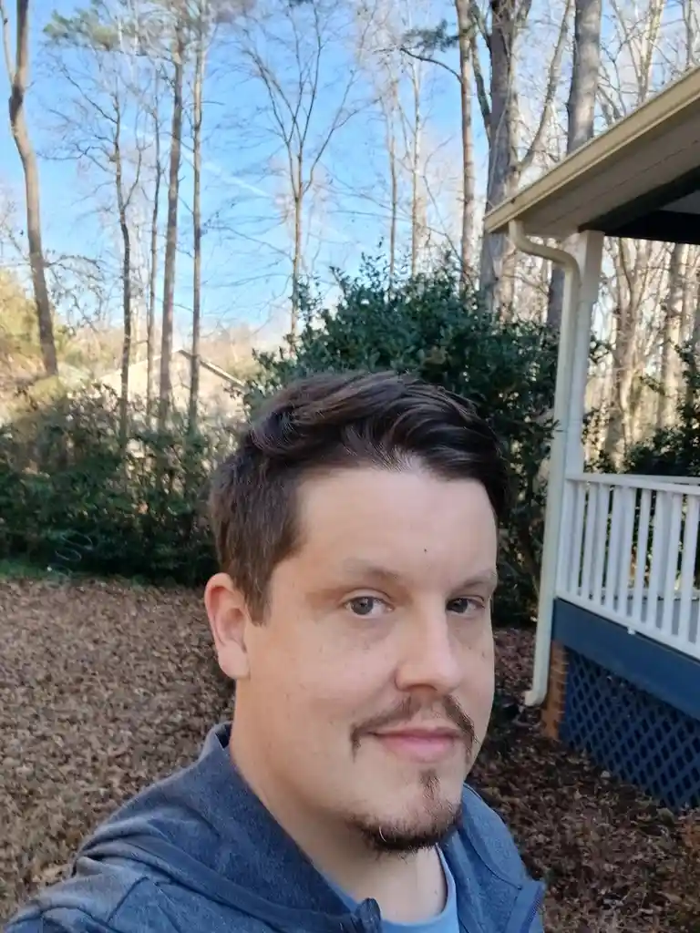 A man with a dark blue hoodie and a goatee stands outdoors in front of a house with a white porch railing and bare trees in…