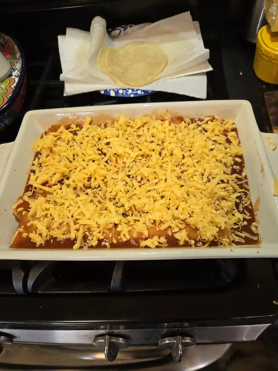 A white baking dish filled with enchiladas covered in a thick red sauce and a generous layer of shredded yellow cheese sits…