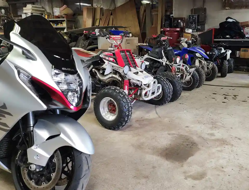 A silver Suzuki motorcycle is prominently featured in the foreground of a cluttered garage, with a row of various ATVs lined…