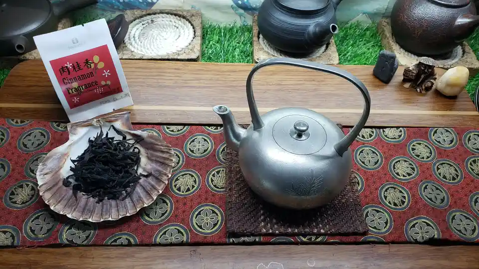 A silver teapot sits on a woven mat atop a red patterned cloth, next to a seashell holding dark tea leaves and a tea bag…