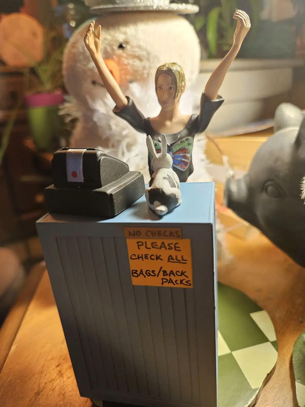 A miniature figurine of a woman with arms raised stands behind a small desk that has a sign reading 'NO CHECKS PLEASE CHECK…