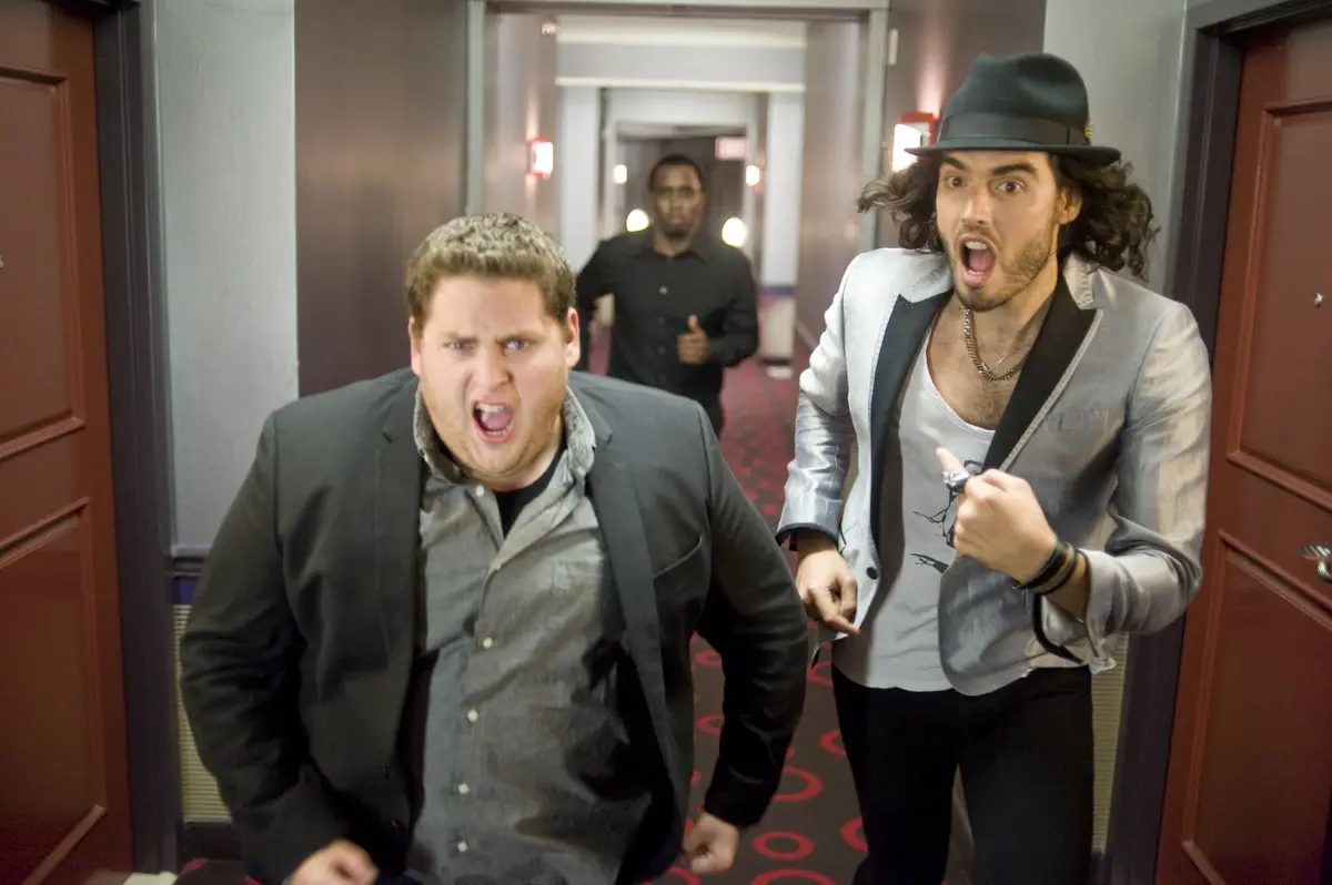 Two men in a hotel hallway, one in a grey suit screaming angrily, the other in a silver jacket and hat pointing with a…
