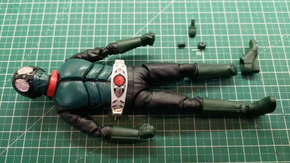A green and black Kamen Rider action figure lies flat on a green cutting mat, with its right arm and left leg detached and…