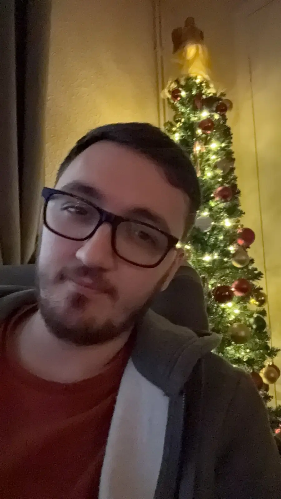 A man with glasses and a beard looks at the camera with a slightly annoyed or unenthusiastic expression, standing in front…