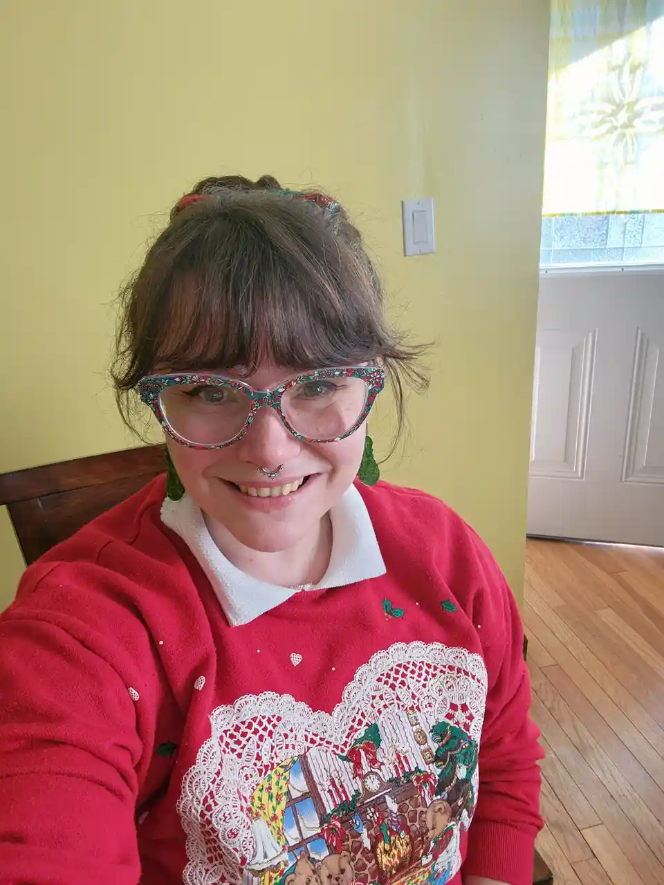 A person with brown hair and bangs smiles at the camera while wearing a red holiday-themed sweater with a white lace heart…