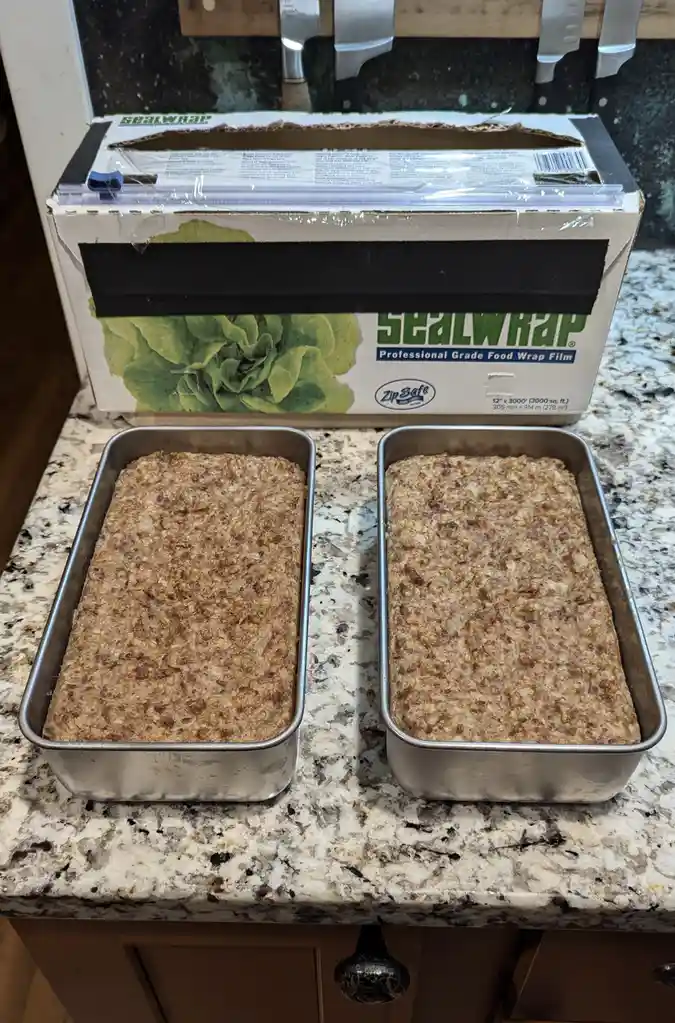 Two rectangular metal baking pans filled with unbaked, textured bread batter sit side-by-side on a speckled granite…