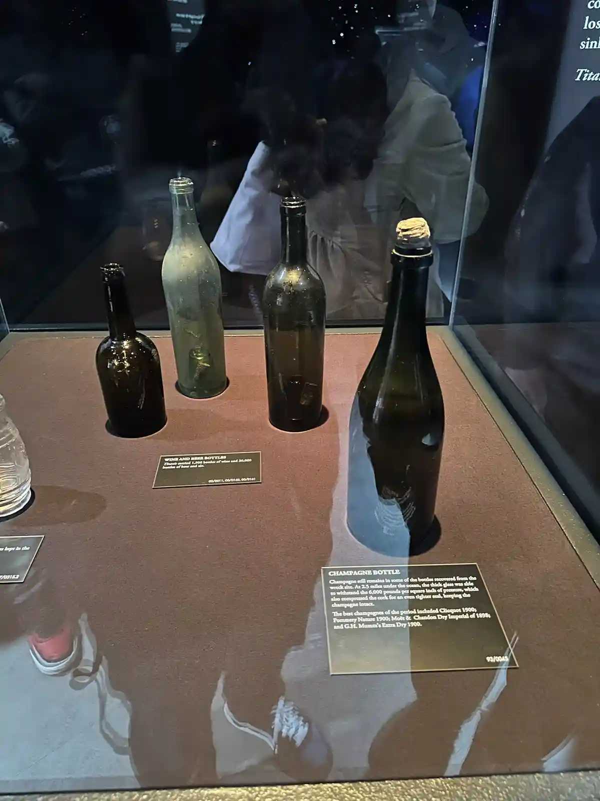 Four vintage glass bottles, including a champagne bottle with a cork, are displayed in a museum case with informational…