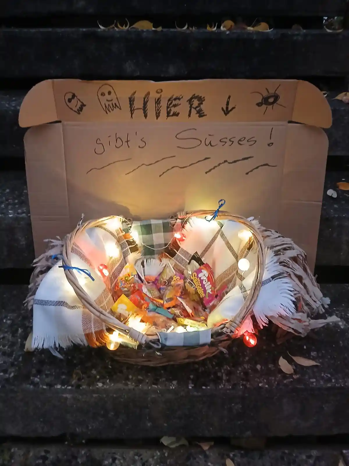 A cardboard box with hand-drawn Halloween faces and the German text 'HIER ↓ gibt's Süsses!' sits behind a decorated wicker…