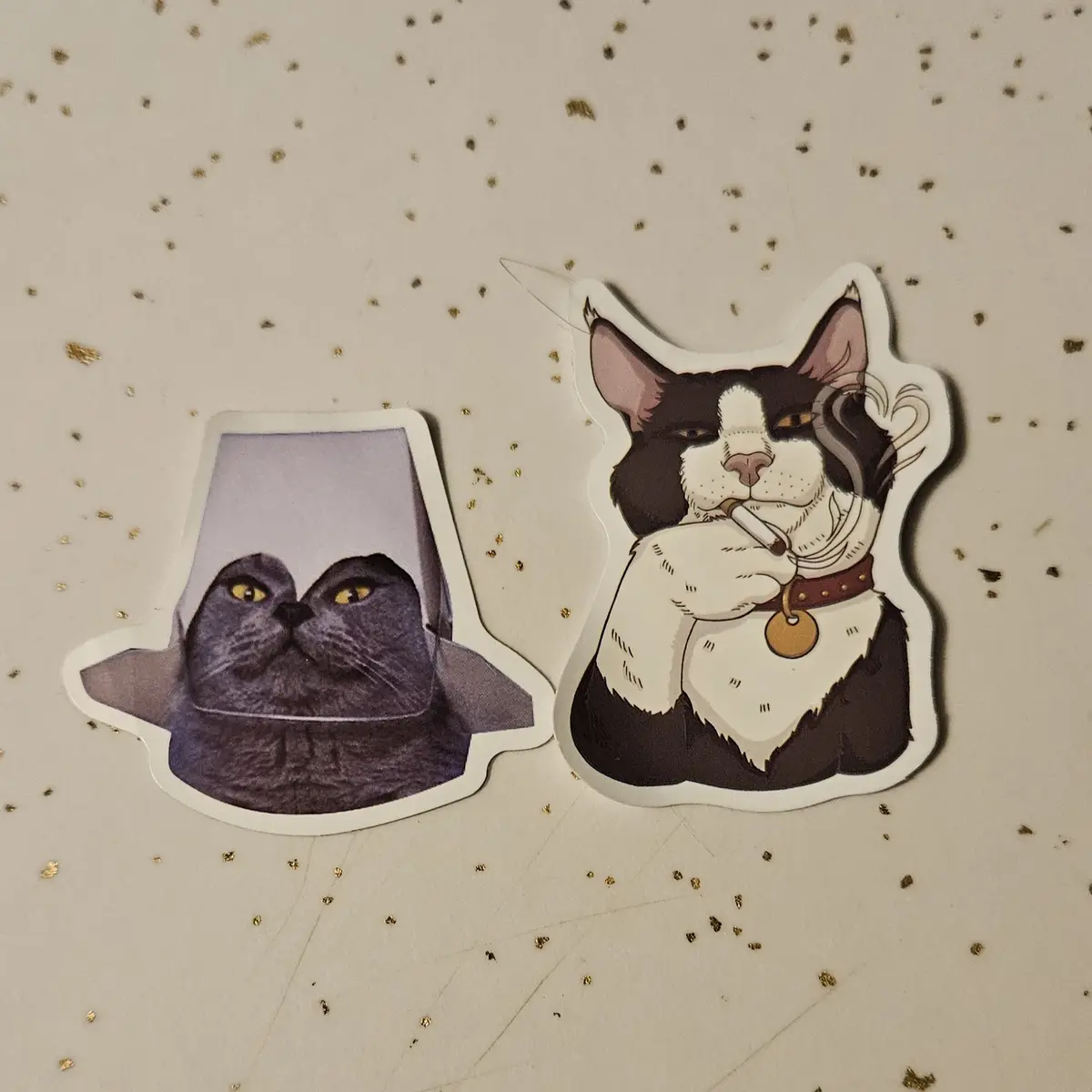 Two cat stickers lie on a glittery surface: one shows a gray cat with yellow eyes peeking out from under a purple…