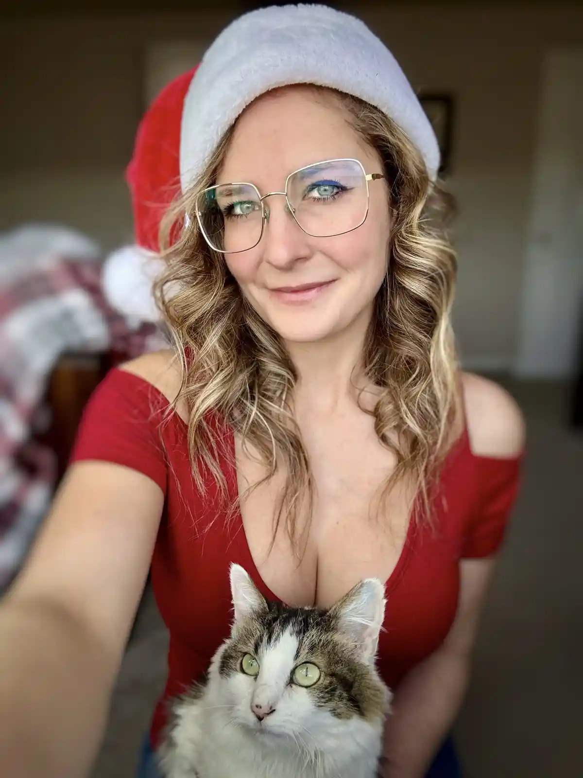 A woman wearing a red and white Santa hat and a red off-the-shoulder top holds a calico cat in her arms while taking a…
