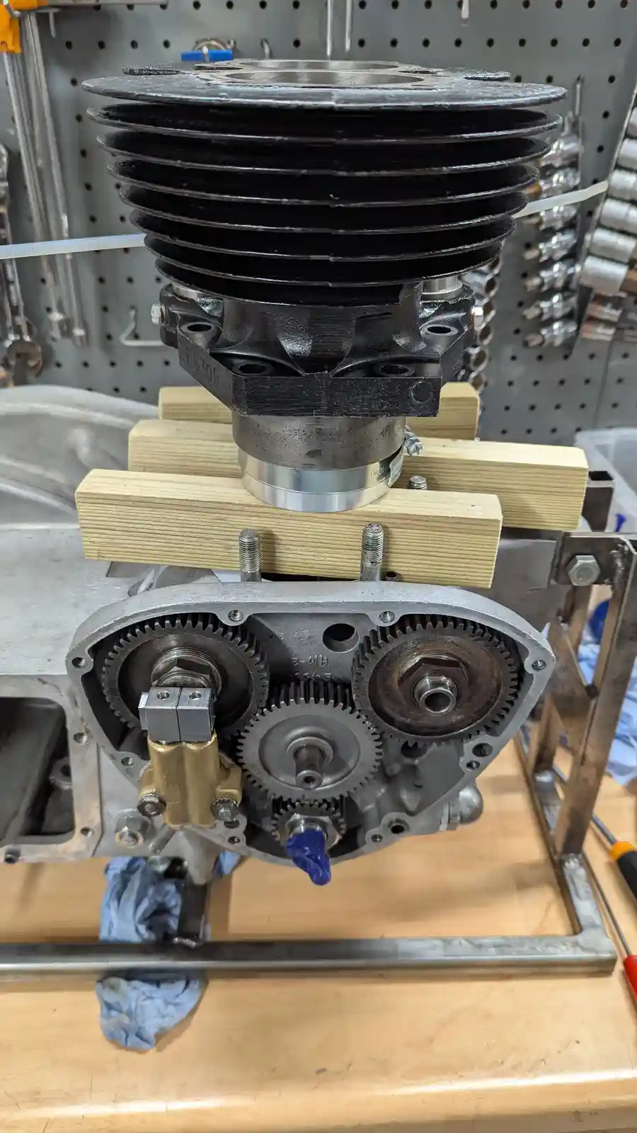 A disassembled engine with its cylinder head and gear train exposed, resting on wooden blocks in a workshop. The overlaid…