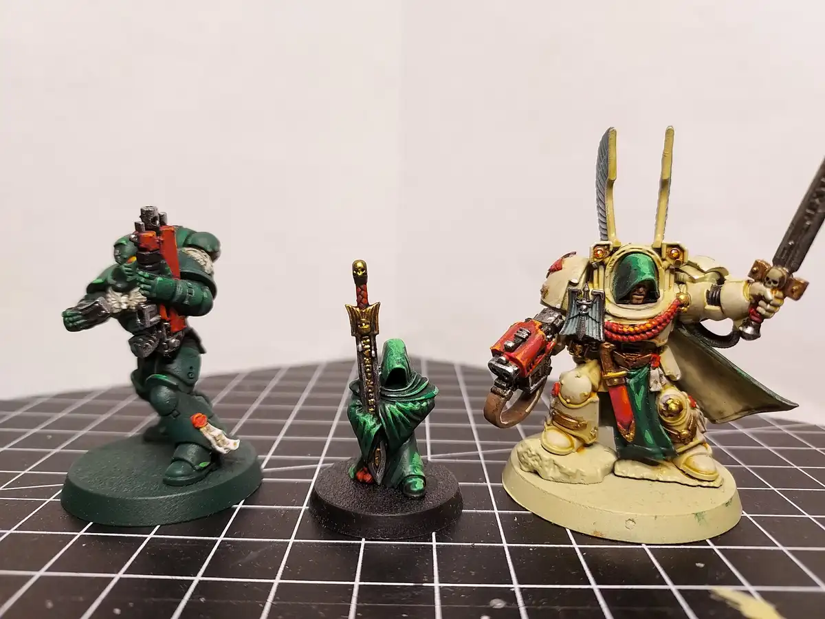 Three painted miniature figurines from Warhammer 40,000 stand on a grid mat: a green-armored Space Marine with a bolter, a…