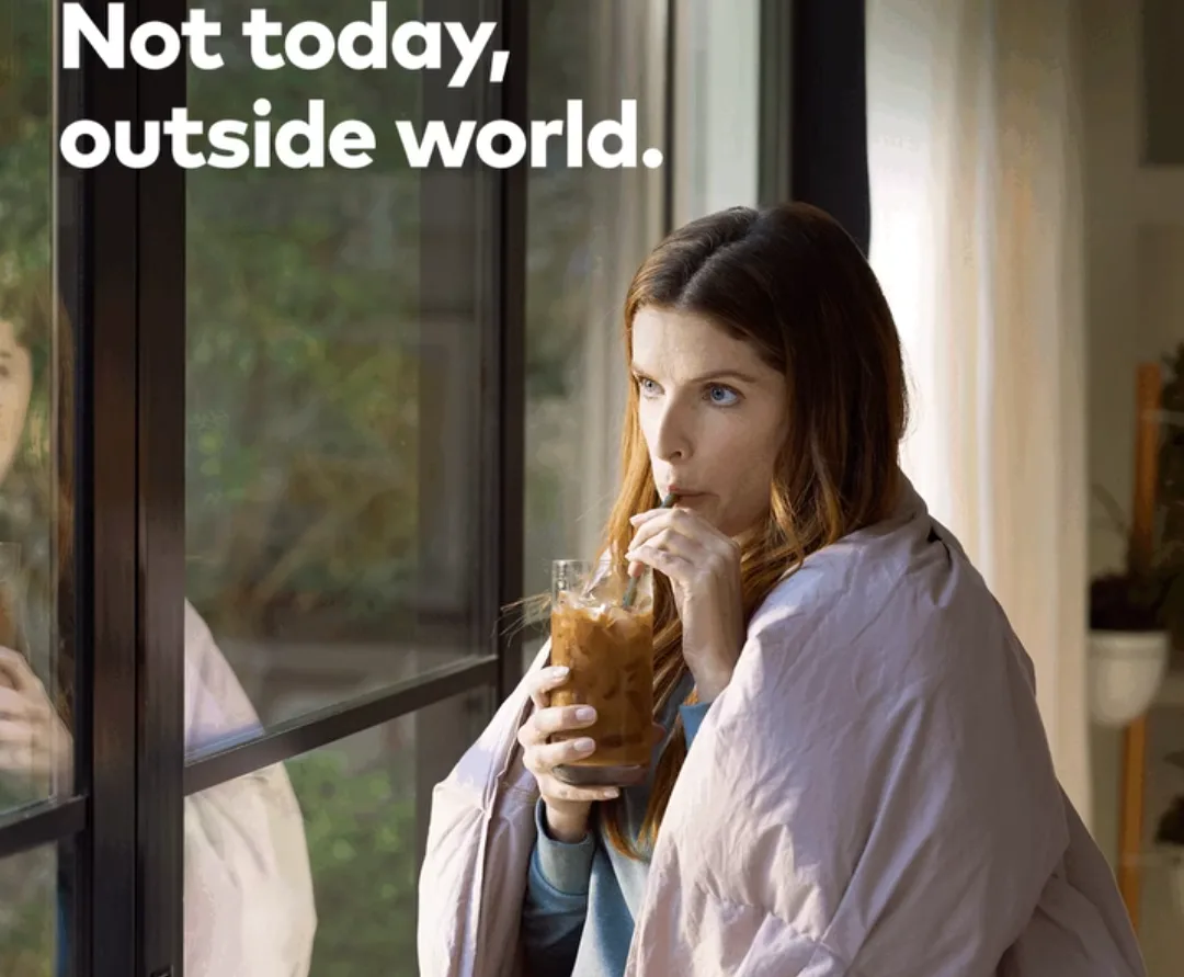 A woman with long brown hair sits by a window, wrapped in a light pink blanket, holding a glass of iced coffee with a straw…