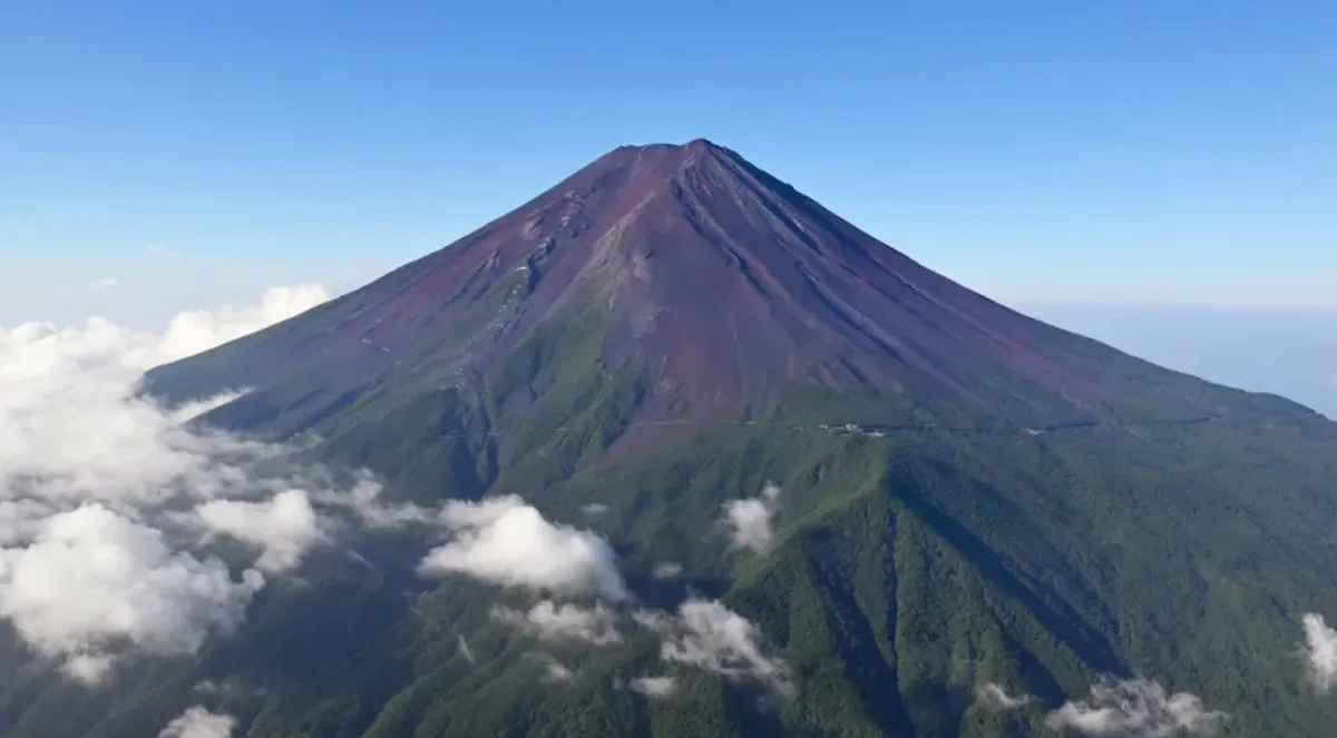 Aerial view of Mount Fuji, Japan, with its iconic conical shape rising above clouds under a clear blue sky. The mountain's…