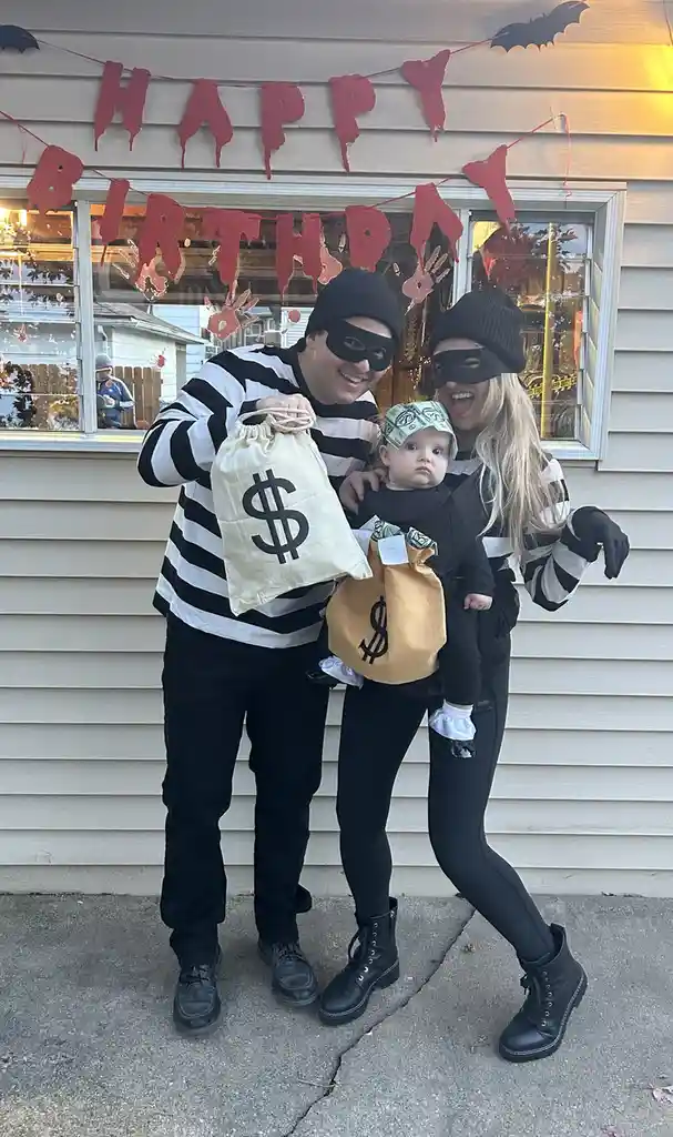 A family of three dressed as bank robbers — a man and woman in black-and-white striped outfits and black masks, holding…