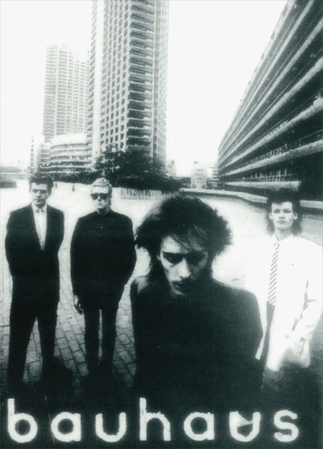 A black-and-white photo of four men standing in an urban setting with tall buildings in the background. The man in the…