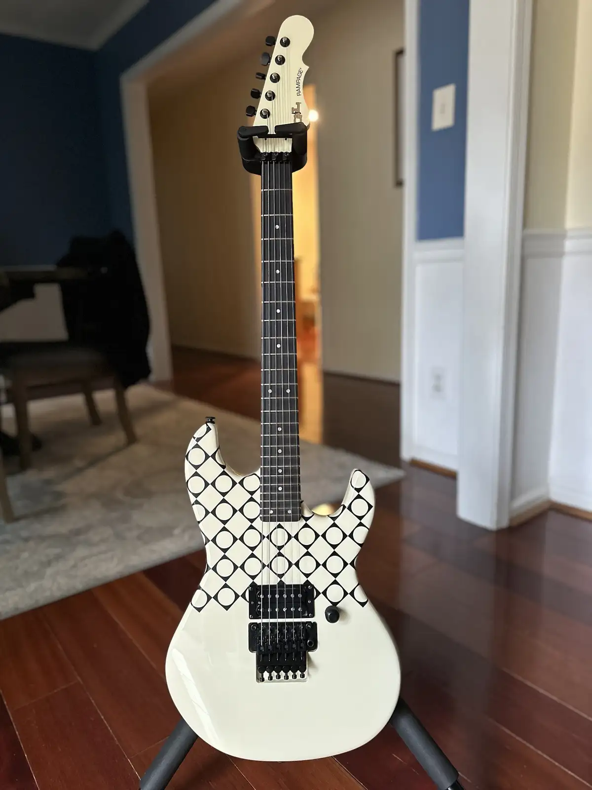 A white electric guitar with a black-and-white checkerboard pattern on its body is standing upright on a stand in a room…