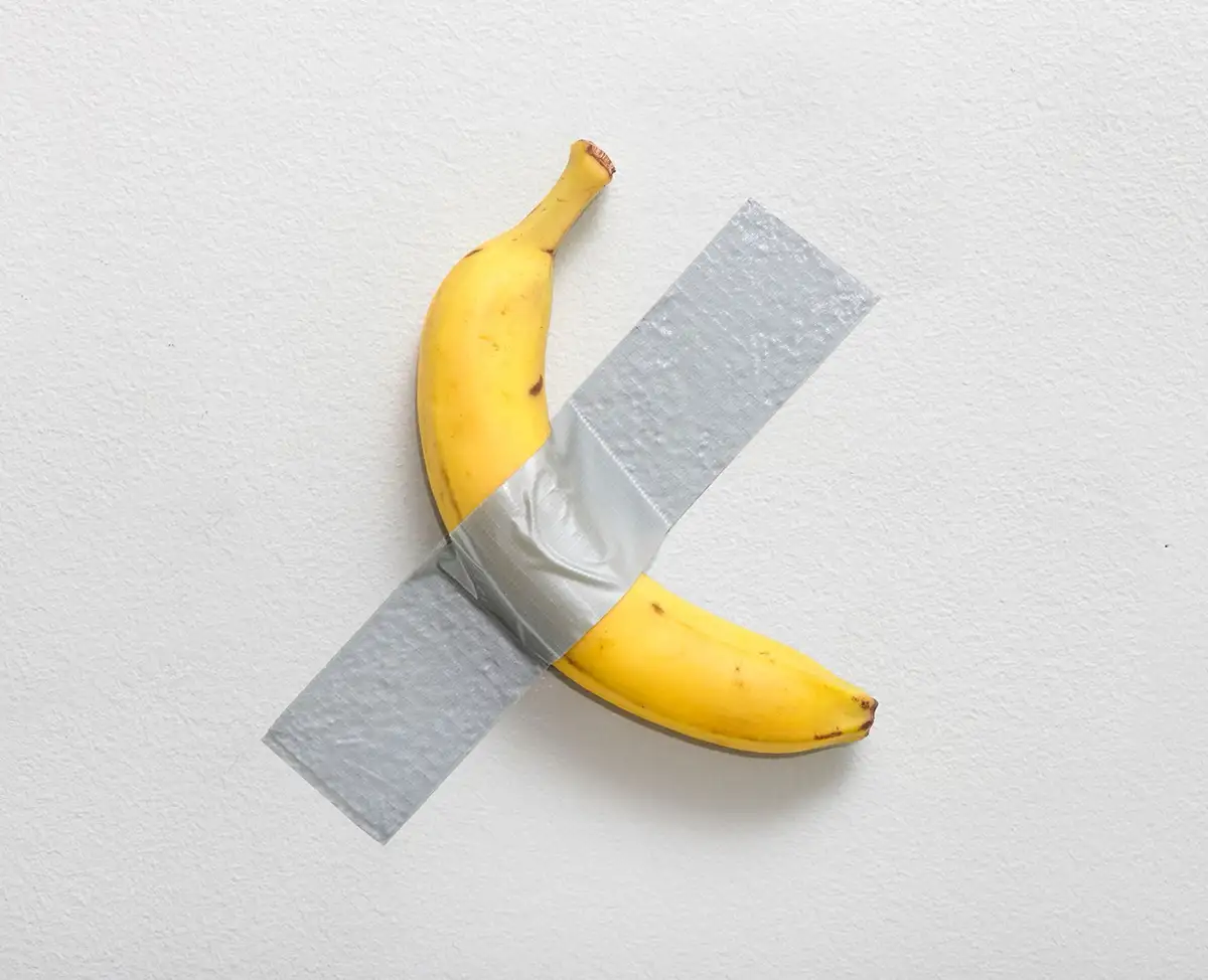 A single yellow banana is taped to a white wall with a large piece of gray duct tape forming an 'X' across its body. The…