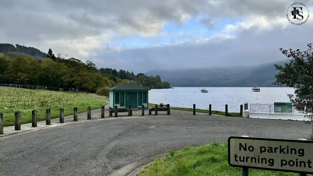 A scenic view of a lake with boats and forested hills under a cloudy sky, featuring a small green building near a road. A…