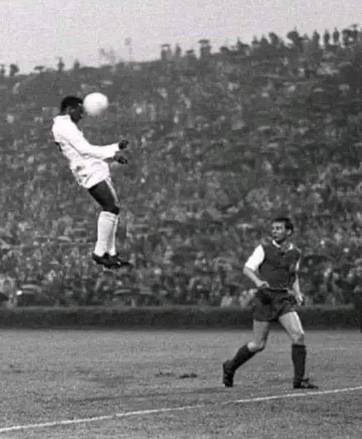 A black-and-white photo shows a soccer player in a white jersey leaping high in the air to head a ball, while another player…