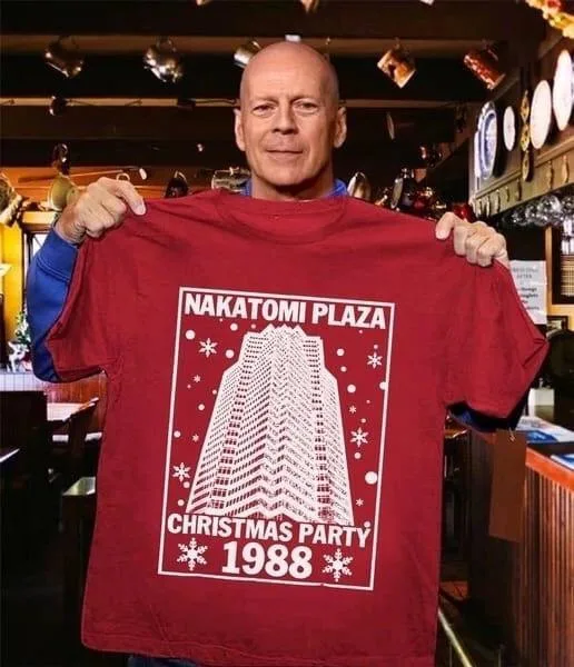Bruce Willis holds up a red t-shirt featuring a white graphic of the Nakatomi Plaza building with snowflakes and the text…