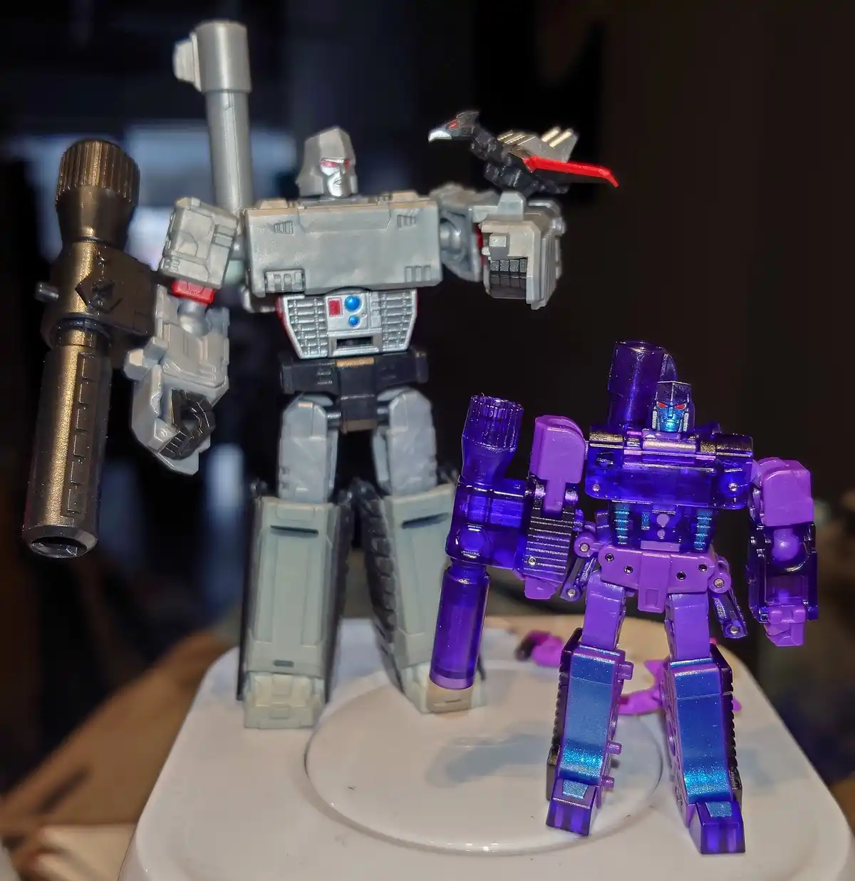 Two Transformers action figures stand side by side on a white display stand; a larger, gray, silver, and black robot with a…