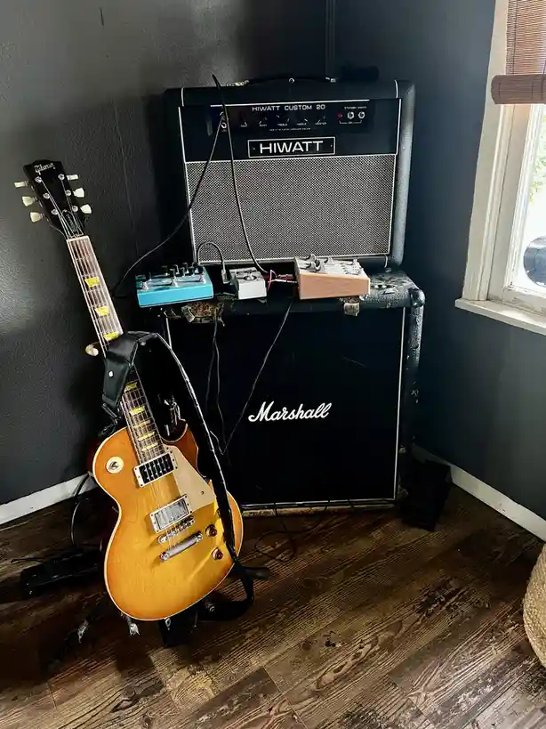 A Gibson electric guitar leans against a wall next to a Hiwatt Custom 20 amplifier head sitting atop a Marshall speaker…