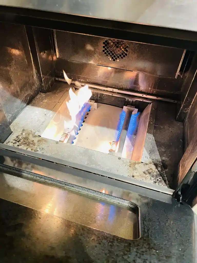 A close-up photo shows the interior of a stainless steel oven with a visible blue and yellow flame erupting from the burner…