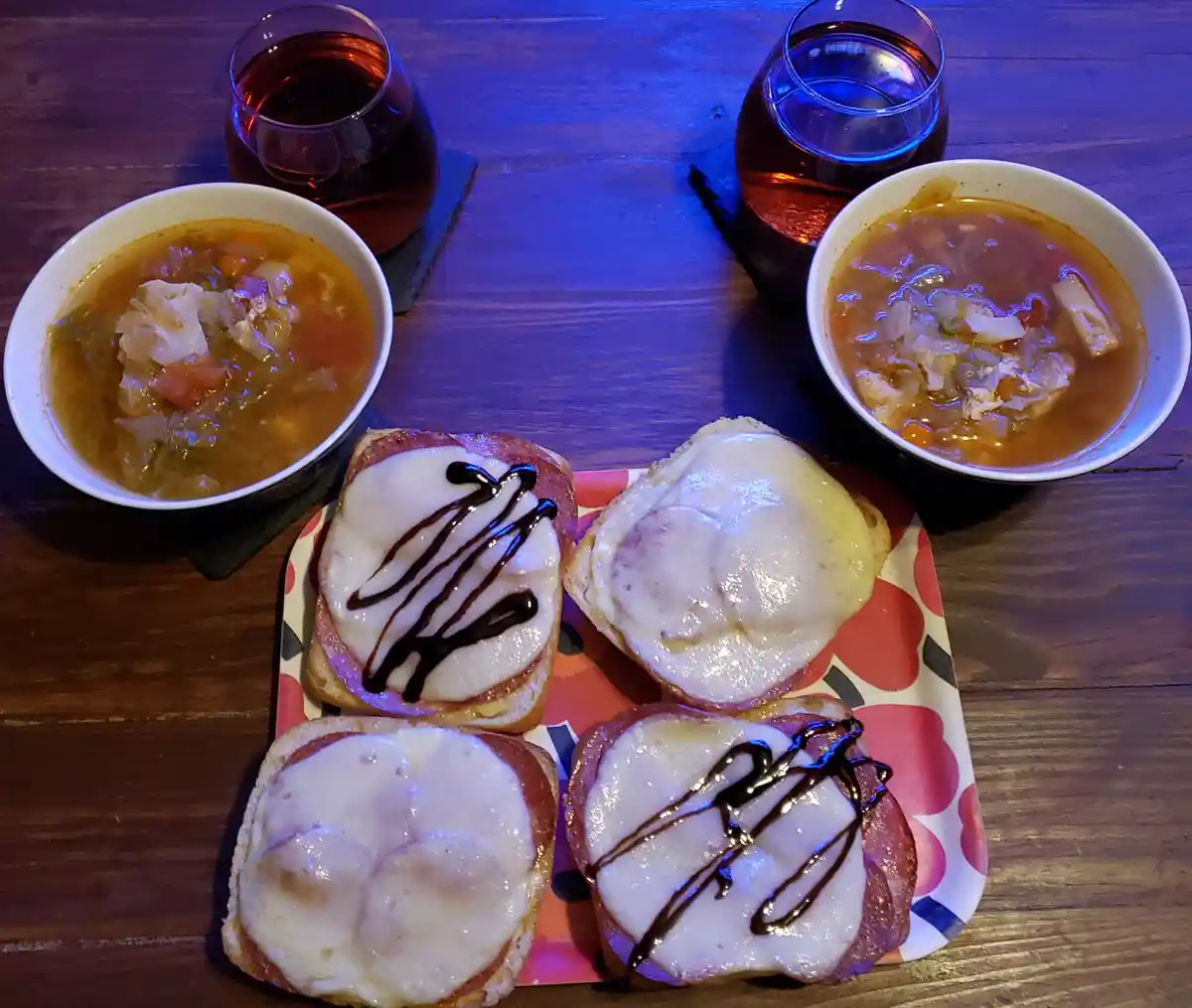 A wooden table holds two bowls of chunky vegetable soup, two glasses of red wine, and a colorful plate with four open-faced…