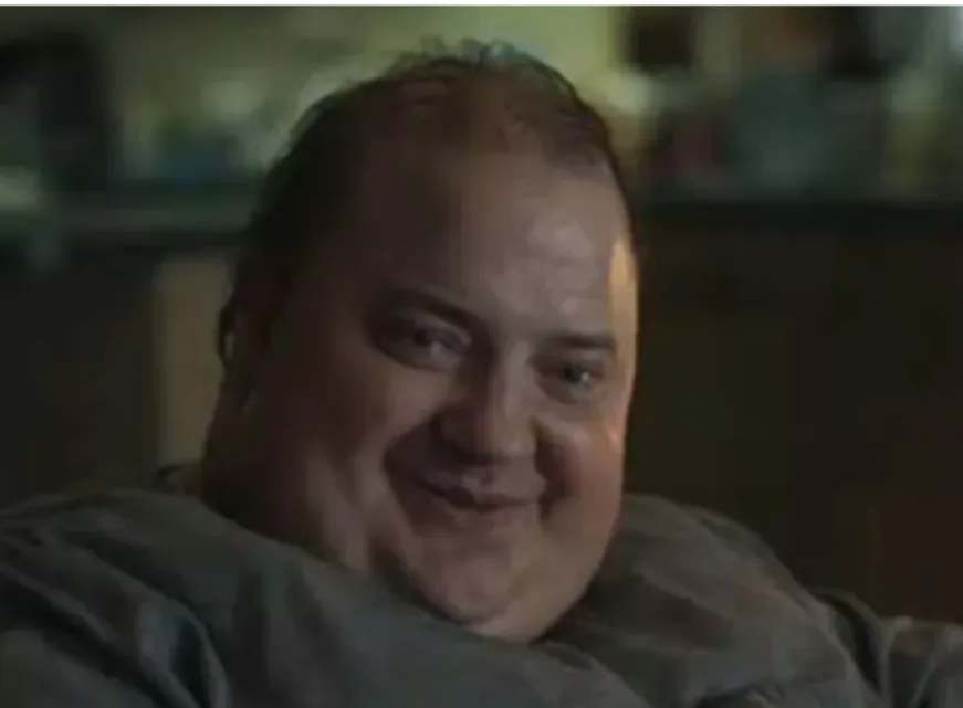 a man with a balding head and a wide, slightly unsettling smile is shown in a dimly lit room, wearing a grey shirt. The…