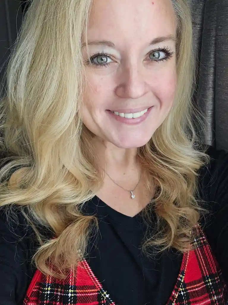 A close-up selfie of a smiling woman with blonde hair and green eyes, wearing a black shirt under a red plaid vest. She has…