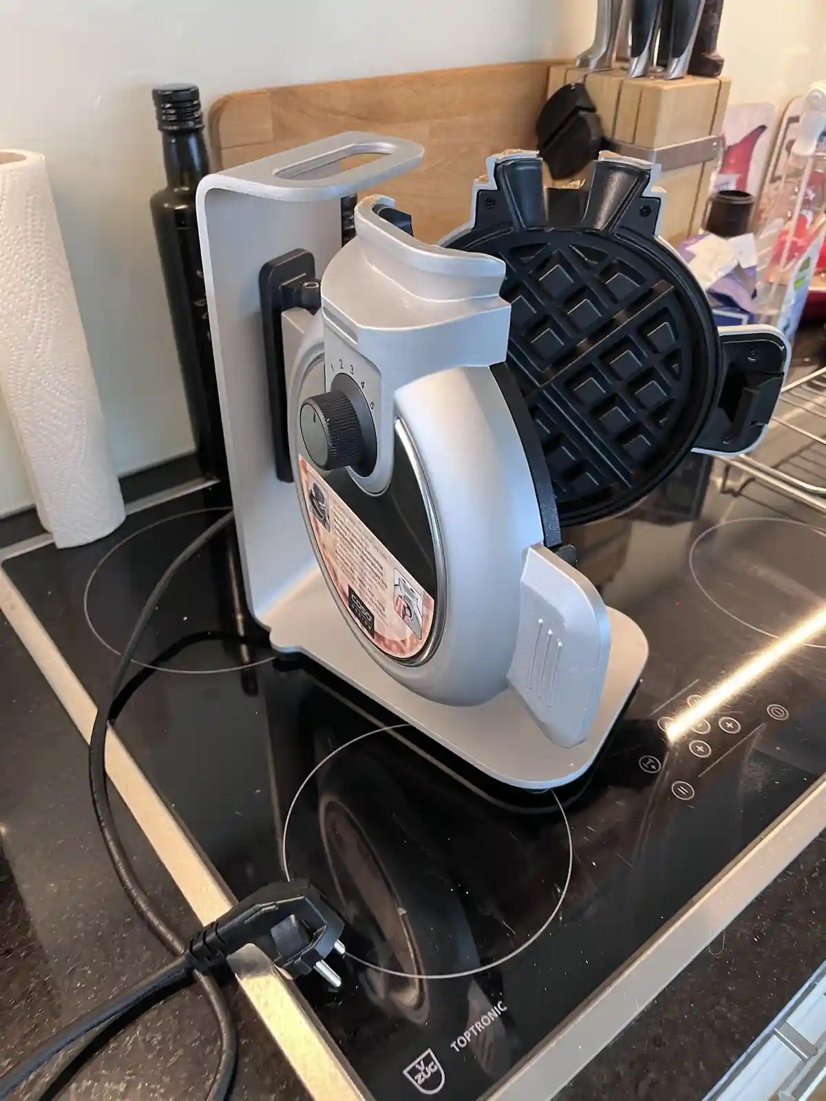 A silver waffle maker sits open on a black glass stovetop, revealing its black grid-patterned cooking plates. The appliance…